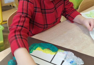 Dziewczynka odbija gąbeczkę z farbą na kolorowym kartonie.