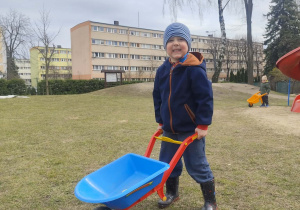 Chłopiec jedzie taczką po ogrodzie.